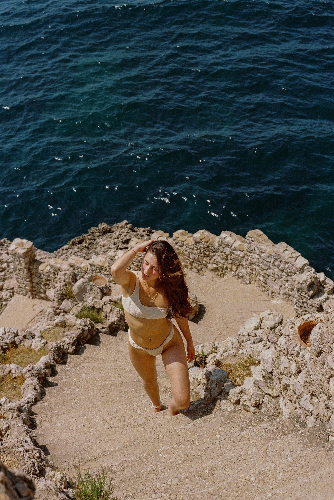 Woman in a bikini standing on stone steps overlooking the ocean.